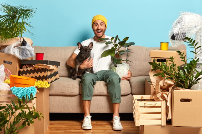 Image of positive fashionable guy rents new flat, lives together with favourite dog, sits on couch, holds houseplant, rejoices cheap loan has many personal stuff and household things. Mortgage concept